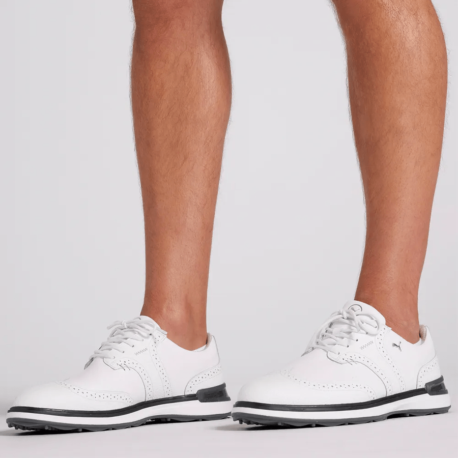 A person wearing PUMA Avant Wingtip Golf Shoes - White with black soles and white laces stands on a light surface, showing only their lower legs and feet.
