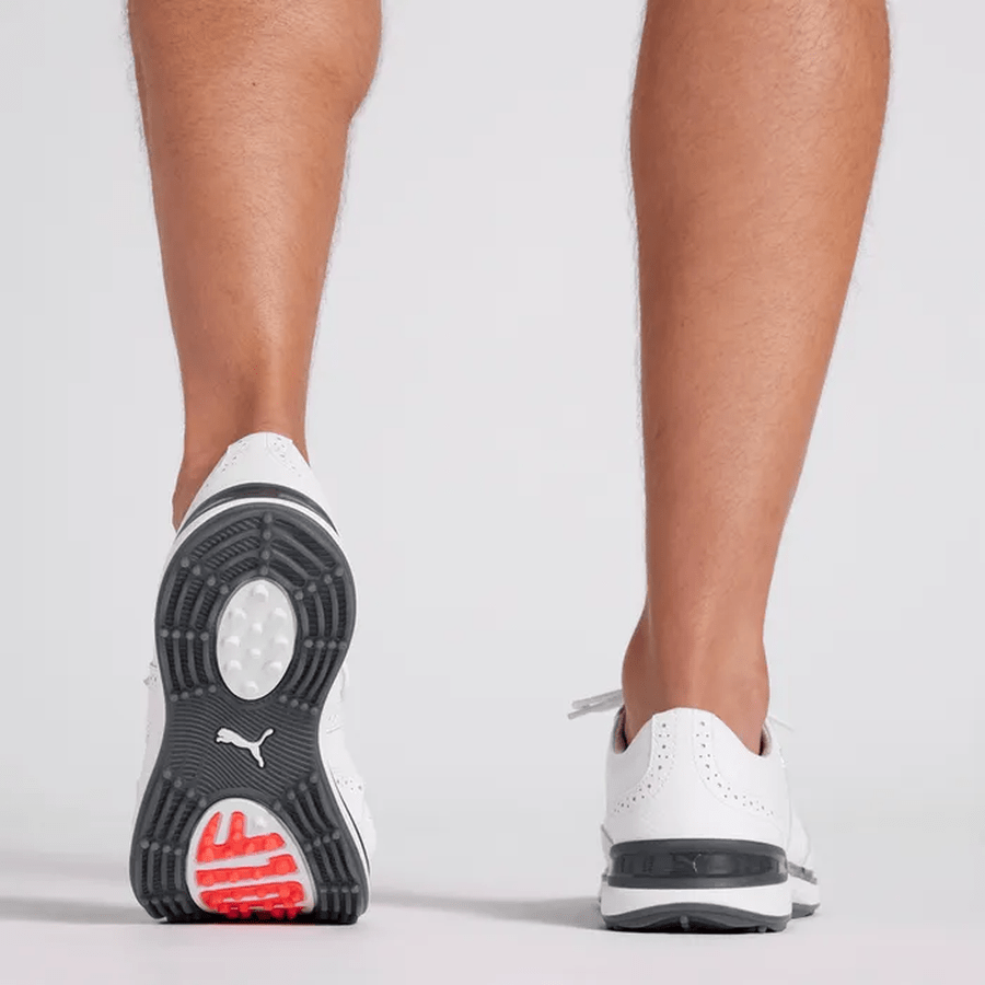 Close-up of a person’s lower legs and feet wearing PUMA Avant Wingtip Golf Shoes - White, featuring black and red details, spikeless design, and advanced radial traction technology, photographed from behind on a plain light background.