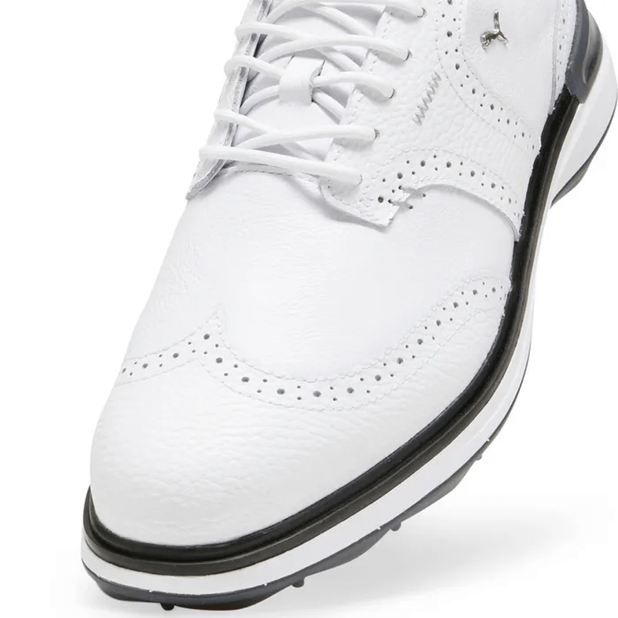 Close-up of PUMA Avant Wingtip Golf Shoes in white with black accents, featuring decorative perforations, white laces, detailed stitching, and radial traction technology for excellent grip and stability on the course.