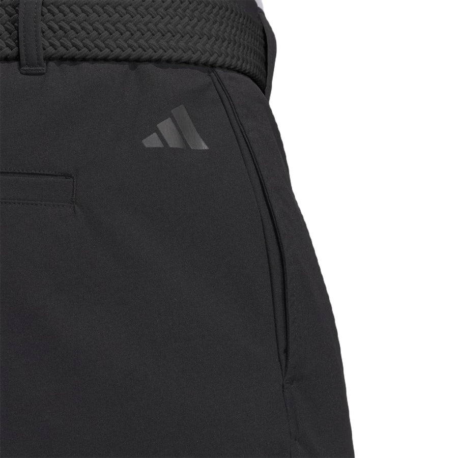 Close-up of a person wearing black Adidas Ultimate365 Chino Pants with a visible pocket and braided black belt. A dark Adidas logo appears above the left pocket. Made with recycled materials for sustainability.