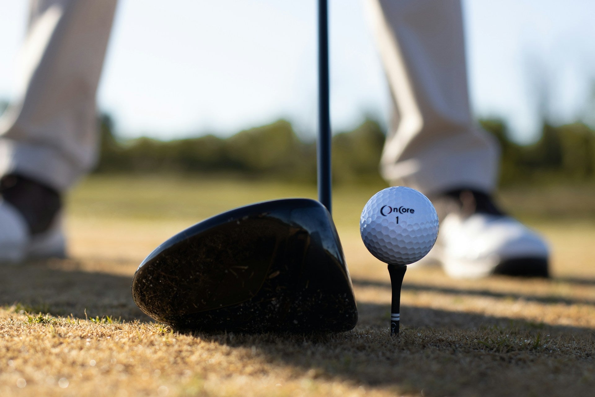 detail of a 7 wood golf club next to a golf ball with the feet of a player visible in the background