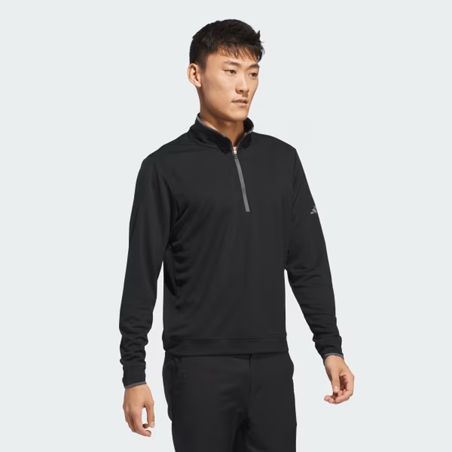 A man in the Adidas Lightweight Half-Zip Top - Final Sale, black in color and made from recycled materials, pairs it with black pants and stands against a plain light background, gazing slightly to the side with a neutral expression.