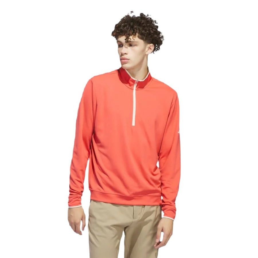 A young man with curly hair wears the Adidas Lightweight Half-Zip Top in bright coral and khaki pants, made from recycled materials, standing against a plain white background.