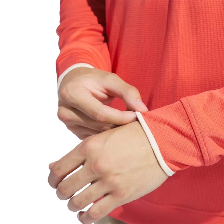 Someone adjusts the cuff of their Adidas Lightweight Half-Zip Top, a long-sleeve coral golf layer made from recycled materials. The background is white.