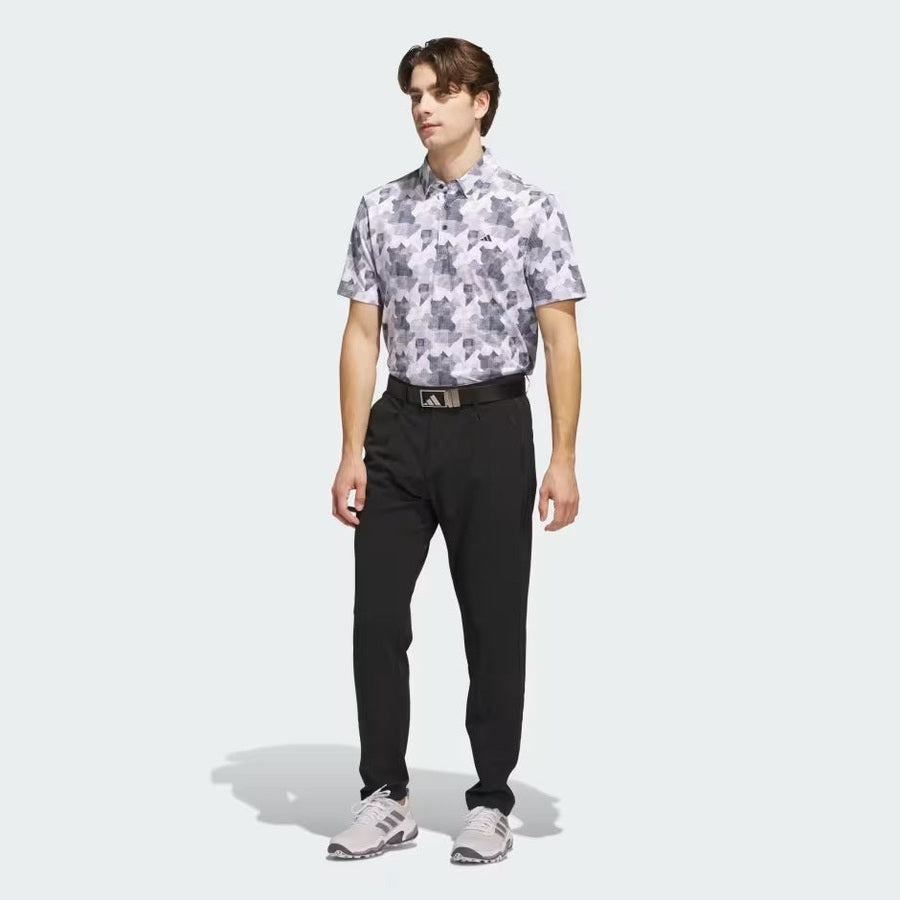 A man wears the Adidas 2025 Go-To Printed Polo Shirt with black pants, a black belt, and light gray sneakers, standing against a plain light gray background.