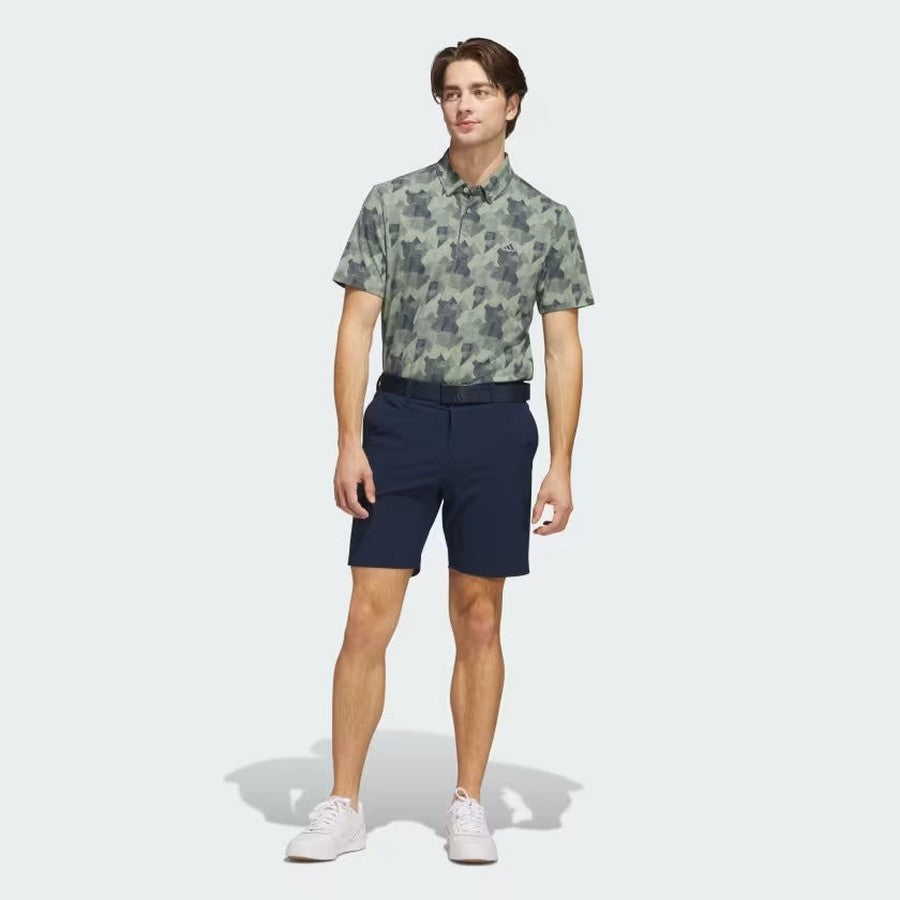 A man in an Adidas 2025 Go-To Printed Polo Shirt (green camo), navy shorts, white sneakers, and a black belt—made from recycled materials—stands against a plain light gray background.